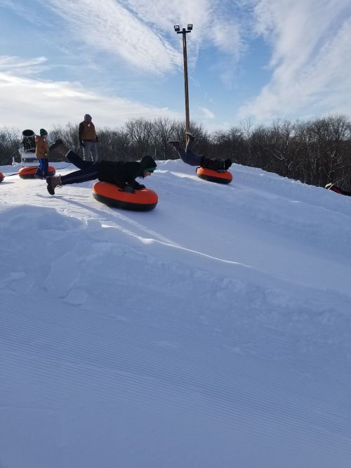 Two people sledding down a snowy hill on orange inner tubes, with a clear blue sky and trees in the background.