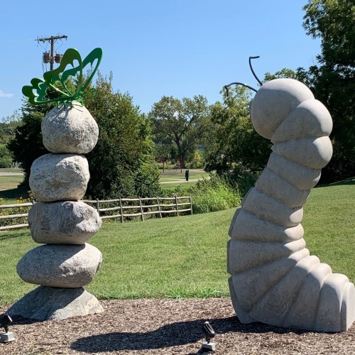 Two whimsical stone sculptures in a park: one resembles a caterpillar, the other a stack of rocks with a butterfly.
