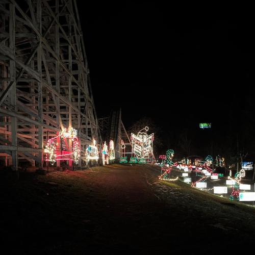 A festive scene at night featuring colorful holiday lights along a path beside a wooden roller coaster.