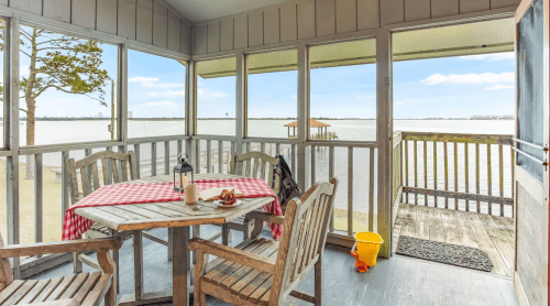 A cozy screened porch with a wooden table and chairs, overlooking a calm water view and a small dock.