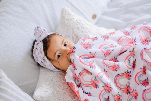 A young child with a floral blanket and headband, lying on a bed surrounded by pillows, looking curiously at the camera.