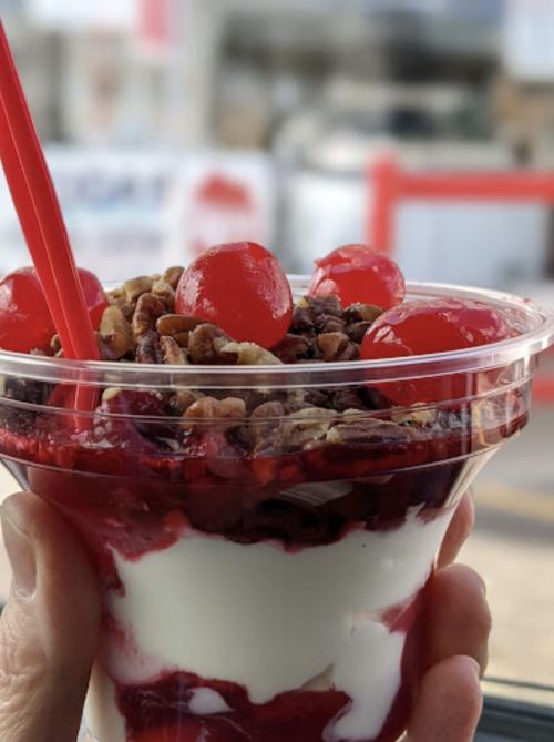 A hand holding a cup of layered dessert with cherries, nuts, and creamy layers, set against a blurred background.