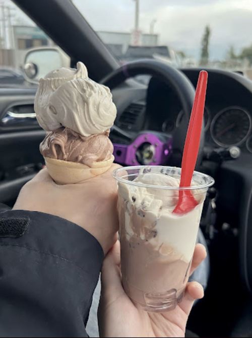 A hand holding a double scoop ice cream cone and a cup of ice cream with a red spoon, inside a car.