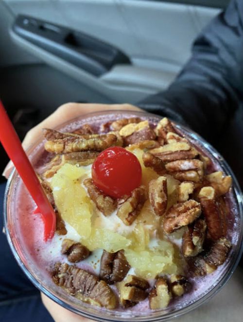 A close-up of a colorful dessert drink topped with nuts, pineapple, and a cherry, held in a hand with a red straw.