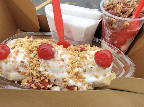 A dessert box with a banana split topped with whipped cream, nuts, and cherries, alongside a cup of strawberry parfait.