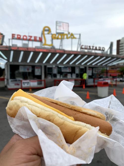A hand holding a hot dog in front of a retro-style frozen custard stand.