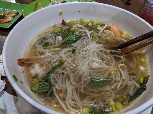 A bowl of noodle soup with shrimp, herbs, and green onions, served with chopsticks on the side.