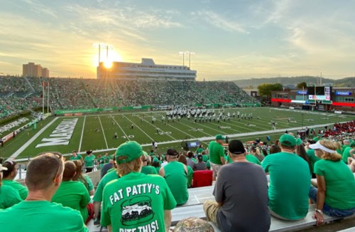 A football game at sunset, with fans in green shirts watching the field and a marching band performing.