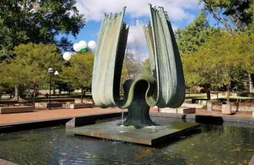 A large, artistic fountain surrounded by trees and greenery, with water cascading from its unique, sculptural design.