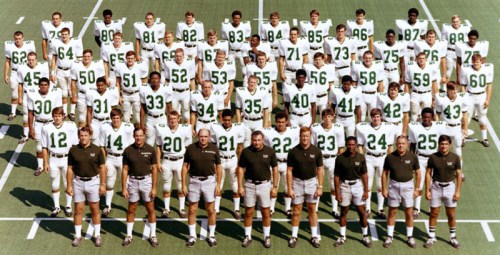 A large group of football players and coaches in green and white uniforms, standing on a field, arranged in rows.