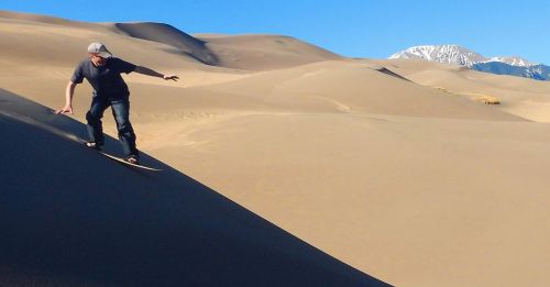A person sandboarding on a dune with mountains in the background under a clear blue sky.