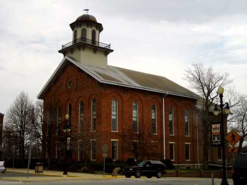 Historic brick building with a cupola, surrounded by trees and parked cars, under a cloudy sky.