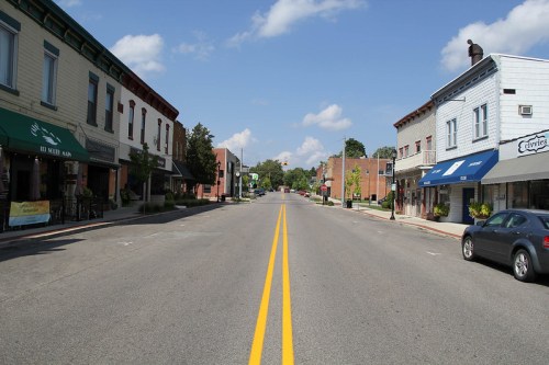A quiet street lined with shops and cafes under a blue sky with fluffy clouds, showcasing a small-town atmosphere.