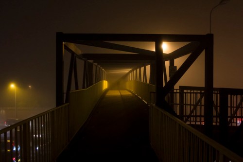 A foggy pedestrian bridge illuminated by streetlights, creating a mysterious and atmospheric scene.