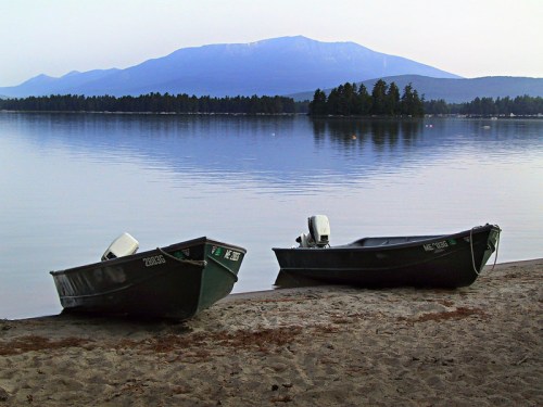Two boats on a sandy shore with a calm lake and mountains in the background, reflecting the serene landscape.