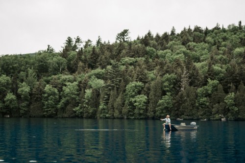 A person fishing from a small boat on a calm lake, surrounded by lush green trees and a cloudy sky.