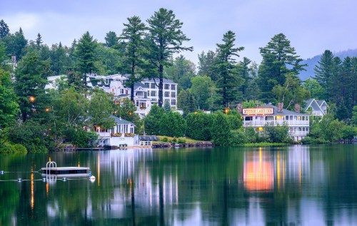 A serene lake scene with lush trees, reflecting buildings, and a calm atmosphere under a cloudy sky.