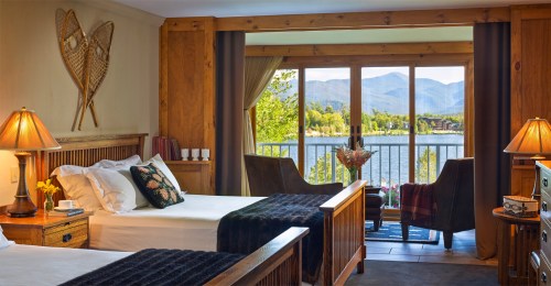 Cozy hotel room with two beds, wooden decor, and a view of mountains and a lake through large windows.
