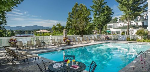 A serene pool area with lounge chairs, surrounded by trees and mountains, overlooking a calm lake on a sunny day.