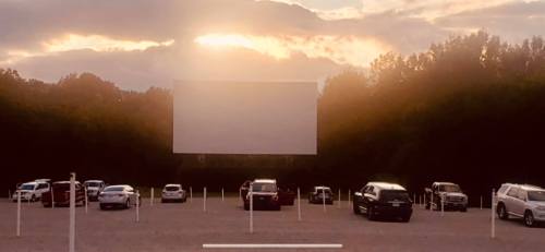 A sunset behind a large blank movie screen at a drive-in theater, with parked cars in the foreground.