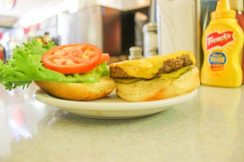 A plate with a burger bun topped with lettuce, tomato, pickles, and a cheese-covered patty, alongside mustard.