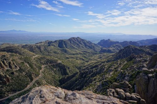 A panoramic view of lush green mountains and valleys under a clear blue sky, with a winding road in the foreground.