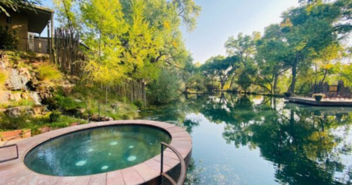 A serene outdoor scene featuring a hot tub beside a tranquil pond surrounded by lush greenery and trees.