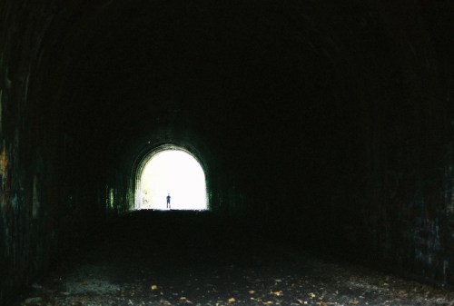 A silhouette of a person stands at the end of a dark tunnel, with bright light illuminating the exit.