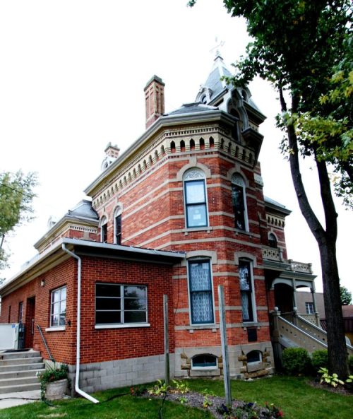 A historic brick house with a turret, ornate details, and a well-maintained garden, surrounded by trees.