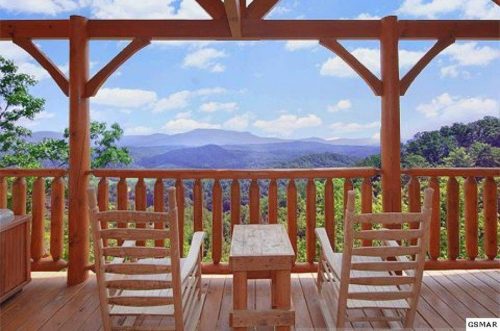 A wooden deck with two rocking chairs overlooks a scenic mountain view under a blue sky with fluffy clouds.