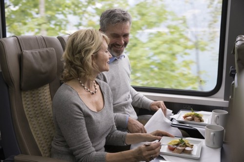 A couple enjoys a meal together on a train, smiling and looking out the window at the scenery.