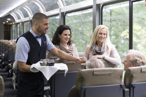 A waiter serves drinks to two women on a train, with passengers seated in the background enjoying the scenic view.