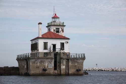 A weathered lighthouse stands on a rocky pier, with a red roof and white walls, surrounded by calm water and a blue sky.