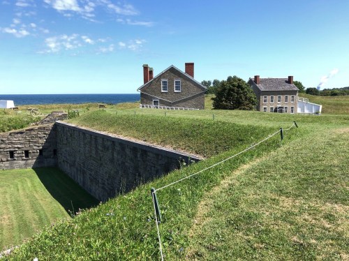 Historic stone buildings on a grassy hill overlooking the ocean under a clear blue sky.