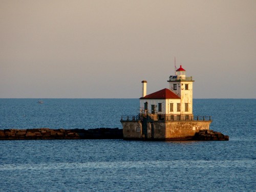 A lighthouse stands on a rocky pier, surrounded by calm water under a clear sky at sunset.