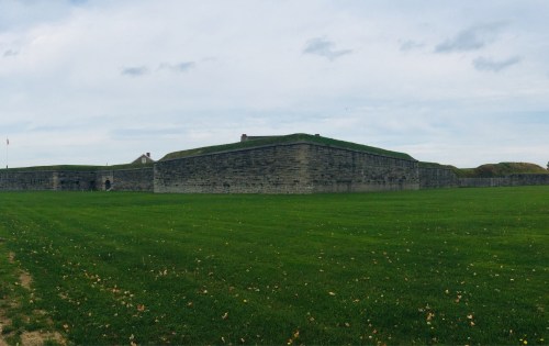A historic stone fort with grassy mounds, surrounded by a green field under a cloudy sky.