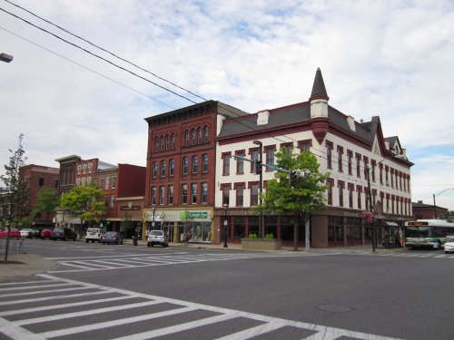 Historic brick buildings line a street corner, with trees and vehicles visible under a cloudy sky.