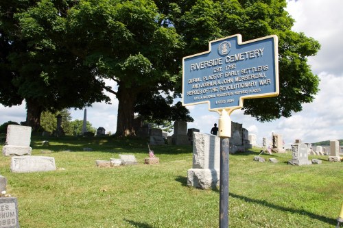 Sign for Riverside Cemetery, established in 1822, with gravestones and trees in the background under a blue sky.