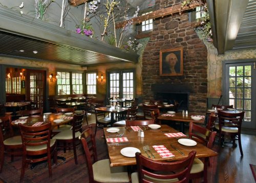 Cozy restaurant interior with wooden tables, chairs, and a stone fireplace, adorned with flowers and a portrait.