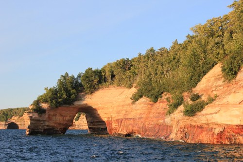 A natural rock arch rises above blue water, surrounded by lush green trees and layered sandstone cliffs.