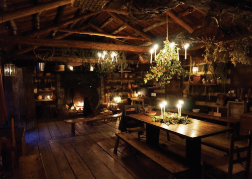 Cozy rustic interior with wooden beams, a fireplace, candles, and a long table surrounded by shelves and greenery.