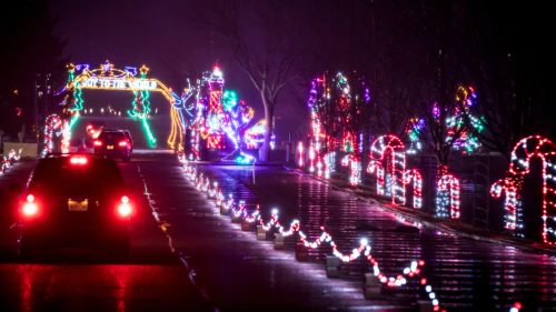 A festive street illuminated with colorful holiday lights, featuring decorations and a car driving through.