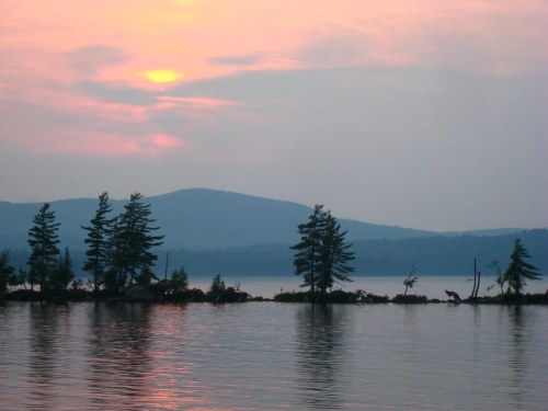 Sunset over a calm lake, with silhouettes of trees and mountains reflected in the water. Soft pink and purple hues in the sky.