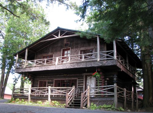 A rustic log cabin with a large balcony, surrounded by trees and featuring a wooden fence in front.