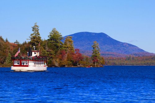 A white boat with passengers sails on a blue lake, surrounded by colorful autumn trees and a distant mountain.