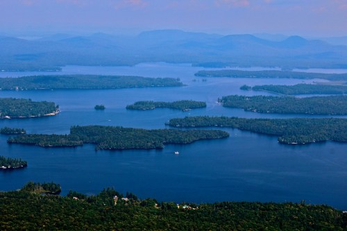 Aerial view of a serene lake surrounded by lush green islands and distant mountains under a clear blue sky.