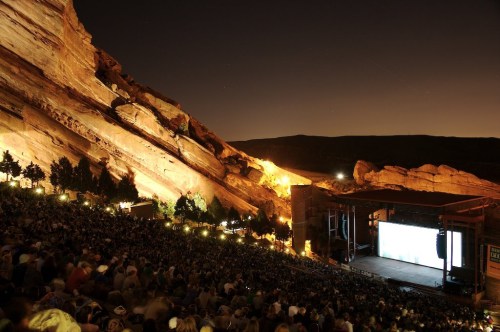 A large crowd gathers at Red Rocks Amphitheatre at night, illuminated by stage lights against a backdrop of rocky cliffs.
