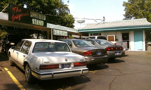 A row of parked cars outside a retro root beer stand with a sign reading "The Original Root Beer Stand."