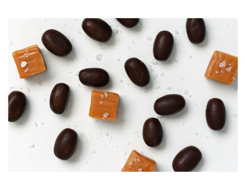 A scattered arrangement of dark chocolate-covered candies and caramel squares with a sprinkle of sea salt on a white background.