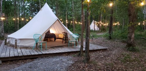 A cozy glamping tent with two teal chairs on a wooden deck, surrounded by trees and string lights in a forest setting.